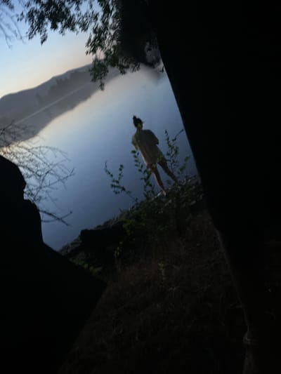 The image depicts a person standing upside down, reflected in a body of water. The person is wearing a light-colored garment and appears to be standing on their hands. The setting is outdoors, with foliage and trees visible on the right side of the frame, suggesting a natural environment. The reflection in the water is clear, mirroring the person and the surrounding greenery. The sky above the reflection is a gradient of blue and white, indicating either dawn or dusk. The overall impression is one of a surreal or artistic composition, playing with perspective and reflection. The image seems to be taken from a low angle, looking upwards towards the person and their reflection. There is no visible text in the image.
