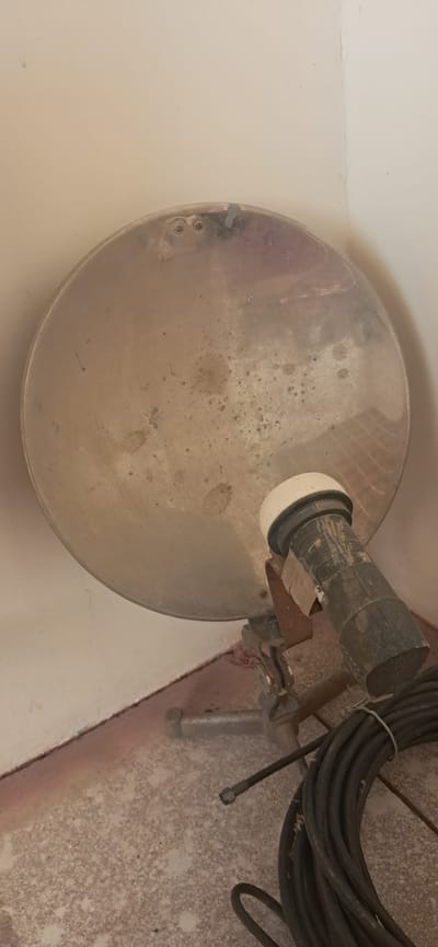 The image shows a satellite dish with a black cable coiled in the foreground, positioned against a cream-colored wall and speckled floor. The satellite dish is round and metallic, with some dirt and discoloration on its surface. A black cylindrical object with a white cap is attached to the dish, likely an LNB. The LNB has some tape wrapped around it. The cable is thick and black, neatly coiled and secured with a white tie. The wall appears to be painted, with some slight discoloration and a visible seam in the corner. The floor is a light-colored, speckled material. The scene is indoors, possibly in a corner of a room or an alcove. There is no direct indication of the specific location in Yazy, Turkmenistan, but the objects suggest a residential or service-related environment.