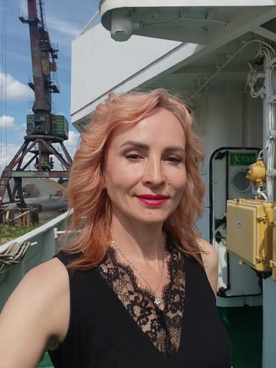 A woman with reddish-blonde, wavy hair, wearing a black top with lace trim and a silver necklace, smiles at the camera. She has on makeup, including bright pink lipstick and eyeshadow. In the background, a large, rust-colored industrial crane stands against a blue sky with scattered clouds. The setting appears to be a port or industrial area, possibly in Vasil'ki, Russia, given the crane and the general industrial aesthetic. The woman is positioned on what seems to be the deck of a ship or a similar elevated platform, indicated by the white railing in the foreground. To her right, a yellow electrical box and a doorway with an "EXIT" sign are visible on a white structure. The overall lighting suggests it is daytime.