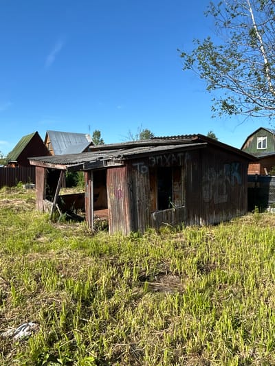 An outdoor, eye-level, medium shot captures an abandoned wooden shed with a corrugated metal roof, situated in an overgrown grassy field under a clear blue sky. The shed is in disrepair, with weathered wooden planks and graffiti spray-painted on its exterior walls. To the left of the shed, another dilapidated structure is visible, and behind it, two houses with prominent roofs can be seen. The overall scene depicts a rural or semi-rural environment, possibly within the city of Слава Снт, Russia, characterized by neglect and overgrowth.
