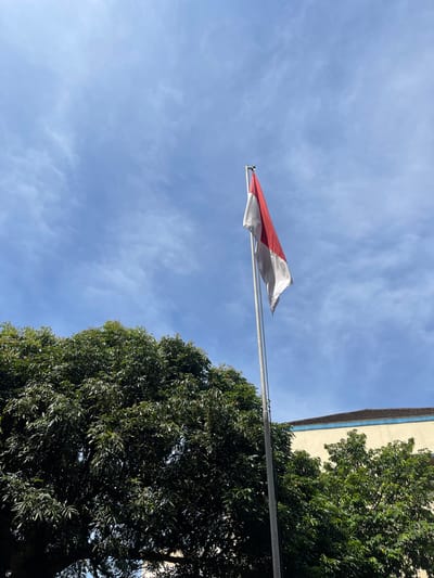 The image shows a view looking up at the Indonesian flag, a red and white bicolor, waving in the wind. The flag is mounted on a silver flagpole. Behind the flag and pole is a bright blue sky with some scattered clouds. To the right of the flag and pole are the green foliage of a large tree, and the corner of a beige building with a dark blue trim. The image might be in the city of Wonosobo, Indonesia.
