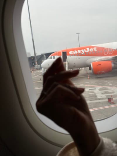 The image captures an exterior view of an airport tarmac from the interior of an aircraft through a passenger window. The scene is located at Lyon-Saint Exupéry Airport (Aéroport de Lyon-Saint Exupéry) in Colombier-Saugnieu, France, under an overcast sky during daylight hours.
In the immediate foreground, a person's right hand and wrist are visible, silhouetted against the brighter exterior. The hand is loosely clasped or resting, with a ring present on the ring finger. The cuff of a light-colored garment is visible around the wrist.
Beyond the window, a large orange and white easyJet Airbus A320 series aircraft is parked on the tarmac. The front section of the aircraft, including its nose, cockpit windows, and the main passenger door (L1), is visible. The passenger door is open, and a set of mobile boarding stairs is positioned at the entrance. The "easyJet" logo is prominently displayed in orange text on the aircraft's white fuselage. The right engine, with its distinctive orange cowling, is also visible. The tarmac features various white and red ground markings, and several orange traffic cones are positioned around the easyJet aircraft. In the background, to the left of the easyJet aircraft's nose, a grey jet bridge structure is discernible, connecting to another terminal or gate. A tall, multi-lamp light pole stands in the mid-background.