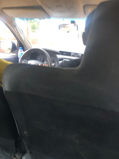 The image captures the interior of a vehicle from a rear passenger's perspective, looking forward through the windshield, located in Mangu Halle, Nigeria. The foreground is dominated by the dark, upholstered back of a front seat, slightly worn. Visible beyond this seat are the steering wheel and a partial view of the dashboard and instrument cluster. A hand, presumed to be the driver's, is positioned on the lower part of the steering wheel. To the left of the steering wheel, a blue-capped plastic water bottle stands upright. The rearview mirror reflects a brightly lit, overexposed outdoor scene. The exterior environment, viewed through the front windshield, is significantly overexposed with visible motion blur, indicating daylight conditions and a vehicle in transit. Faint outlines of trees or vegetation are discernible on the left side of the exterior view. Overhead, the vehicle's sun visors and a roof console are visible. A yellow fabric item is partially visible on the far left of the frame.