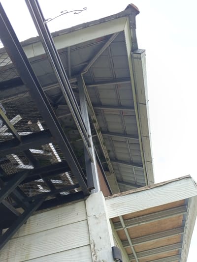 The scene is an upward-angled view of overlapping architectural structures against a bright, overcast sky in Man Gyat, Myanmar. In the foreground, a robust, dark-colored metal framework with an integrated mesh grid extends diagonally upwards from the left. Beneath this, a light-colored, horizontally paneled wall or fascia, showing signs of dirt and discoloration, is visible. A thick, light-colored vertical concrete or painted post supports the metal framework, with a small, dark rectangular object, possibly a light fixture, attached to it. Green vines with broad leaves are observed growing through the metal framework and along an interior column.
To the right and further back, the underside of a roof structure is visible, composed of light-colored metal panels supported by a grid of light-colored metal beams. Its eave features a light-colored fascia board, and a single terracotta-colored roof tile is visible at the top right corner. A decorative, scrolled metal hook is affixed to the fascia board near the top center. In the bottom right, a lower roof or awning is visible from below, exposing light-colored plywood or particle board panels supported by a light-colored metal framework, with a matching horizontally paneled fascia. No individuals or specific activities are present in the image.