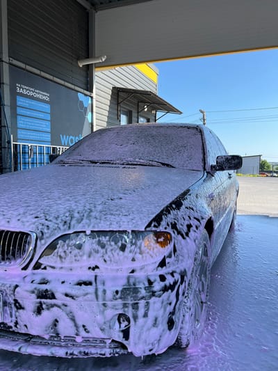 An eye-level, medium shot captures a dark-colored car covered in pink foam at a car wash facility. The car, a sedan, is positioned under a covered bay, with part of the roof and wall of the bay visible at the top of the frame. The car's headlights and front grille are partially obscured by the foam. The ground around the car is wet and soapy. In the background, a building with a grey, corrugated facade and a yellow awning is visible, along with a bright blue sky. A sign on the wall behind the car displays text in Ukrainian. This scene is located at the car wash facility in the city of Velyki Hai, Ukraine.