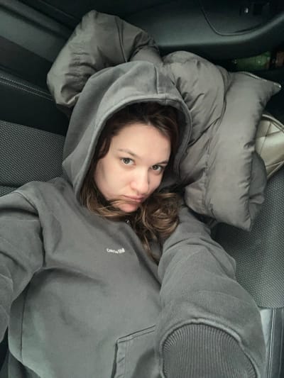 A female individual is depicted in the interior of a vehicle, lying on her back, angled slightly towards the camera. She has light brown, wavy hair and is wearing a dark grey hooded sweatshirt with the embroidered text "Only the Shit" on the left chest area. Her expression features a slight pout, and she looks directly into the camera. Her right arm, mostly obscured, appears extended to capture the image. A grey puffer jacket or blanket is draped over her left side and extends towards the upper right of the frame. To the right of the individual, a light-colored quilted bag is visible, partially obscured by the puffer jacket, and a green bottle is partially visible further back. The vehicle's interior features dark grey patterned fabric on the seat and dark plastic paneling. The specific location within Stara Pazova Municipality, Serbia, is not discernible from the vehicle interior depicted.