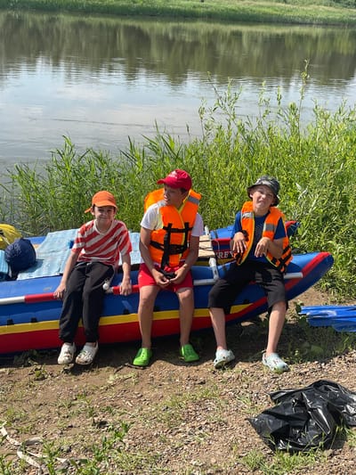 Three young boys, wearing life jackets and casual clothing, are sitting on the edge of a deflated blue and red raft on the muddy bank of a wide, calm river. The boy on the left is wearing an orange cap and a striped t-shirt. The middle boy is wearing a red cap and orange life jacket over a white t-shirt and red shorts. The boy on the right is wearing a camouflage bucket hat and an orange life jacket over a blue t-shirt and black shorts. The river reflects the green foliage on the opposite bank. Tall green grass grows along the bank behind the raft. A dark blue rolled-up sleeping bag and yellow bag are on the raft to the left. A crumpled black plastic bag is on the ground to the right of the raft. The scene appears to be in a natural setting, likely a park or recreational area, near the village of Nezhinka, Russia.