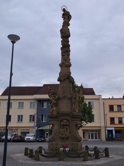 This media file depicts a stone column with a statue of a woman on top, likely a religious figure, adorned with a halo. The column itself is ornate, featuring carvings of figures and cloud-like motifs. Two smaller statues of men in cloaks and halos, holding shields and flags, are positioned on opposite sides of the column's base. At the very bottom of the column, a carved panel shows a group of people. The column stands in a paved public square, surrounded by a low stone barrier with chains.

In the background, a row of buildings lines the square. The central building appears to be a hotel, indicated by a prominent "HOTEL" sign. To the left of the hotel, there are buildings with light yellow and blue facades featuring multiple windows. To the right of the hotel, a building with an orange facade has shops at street level, one of which has a sign reading "ODEVY &..." (likely "Odevy", meaning "clothing" in Czech). Parked cars are visible in front of some of the buildings, including a white van, a black sedan, and a white SUV. A lamppost with a distinctive wide shade stands to the left of the column.

The sky is overcast and appears to be a cloudy day. The overall atmosphere is subdued, and the time of day is likely daytime, given the lighting. The scene appears to be a typical European town square, with architectural styles suggesting a Central European location. No specific event or activity is clearly depicted, but the setting suggests a public space often used for gatherings or as a central point in a town. The scene conveys a sense of historical and cultural significance due to the prominent monument.