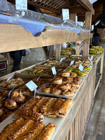 This image captures an interior view of a bakery or food stall in Tsovagyugh, Armenia. The foreground features a multi-tiered wooden display counter filled with an extensive assortment of baked goods. Trays hold various pastries, including golden-brown puff pastry items, spiral-shaped pastries, croissants, and elongated rolls. Some items are identified by white price tags, displaying prices such as "100 դր" (AMD), "330 դր", and "350 դր" for a "PROFESSIONAL ROLL WITH NUT". Several food items on the upper tier are covered with clear plastic dome lids. In the midground, additional displays showcase items like green-topped pastries, and a container holds colorful lollipops on sticks. The display structure is constructed from light-colored wooden planks and beams. The floor is covered with light brown, plank-like material. In the background, a large, light-colored conical stone or clay oven, characteristic of a traditional tonir, is prominent on the left. To the right, a person in dark pants and a jacket is partially visible walking, while another individual wearing a dark jacket stands with their back to the camera. The scene suggests an active retail environment for baked goods.