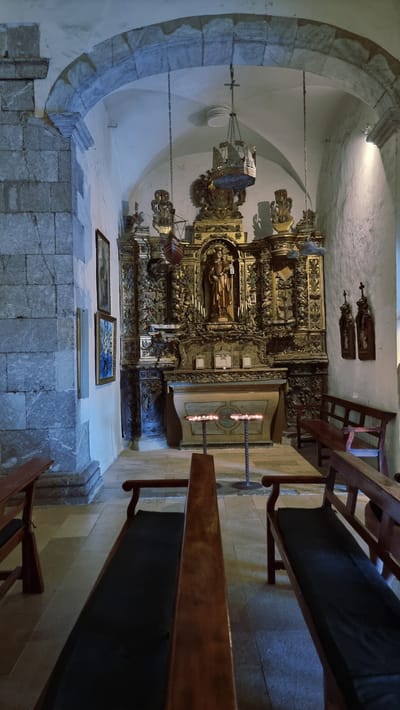 The image depicts the interior of a church in Cadaqués, Spain, focusing on the altar and the immediate surroundings. The scene is dimly lit, with light emanating from candles on the altar and from a light source above and to the right, illuminating the textured white wall. A large, ornate golden altarpiece dominates the background, featuring a statue of a religious figure holding a child in its center. Flanking the central altarpiece are smaller, detailed decorative elements. In front of the altarpiece, there is a table with two sets of candles burning on stands. The floor is tiled with large, light-colored rectangular tiles. To the left of the altar, a stone column and wall are visible, with two framed religious paintings hanging on the wall. In the foreground, wooden pews with dark cushions are arranged facing the altar, with the back of one pew partially obscuring the view of another. The architectural style suggests an older building, with arched stone structures visible at the top of the frame. There are no people or animals present in the image.