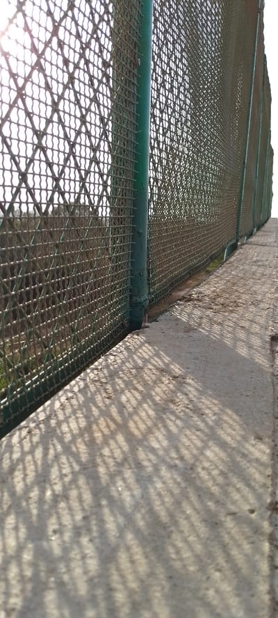 A close-up shot showcases a green metal chain-link fence with a vertical support pole. The fence casts a patterned shadow onto a concrete surface below. Beyond the fence, glimpses of a structure and vegetation are visible. The overall perspective gives a sense of depth. The sun seems to be casting its light from the left, enhancing the shadows.
