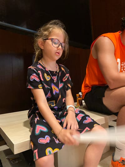 A child with blonde hair and glasses is sitting on a light-colored surface next to a person wearing an orange shirt and black shorts. The child is wearing a black dress with colorful heart patterns and is holding a white object. There is a chocolate bar on the surface next to them. The location is Marina Maroma, Mexico.