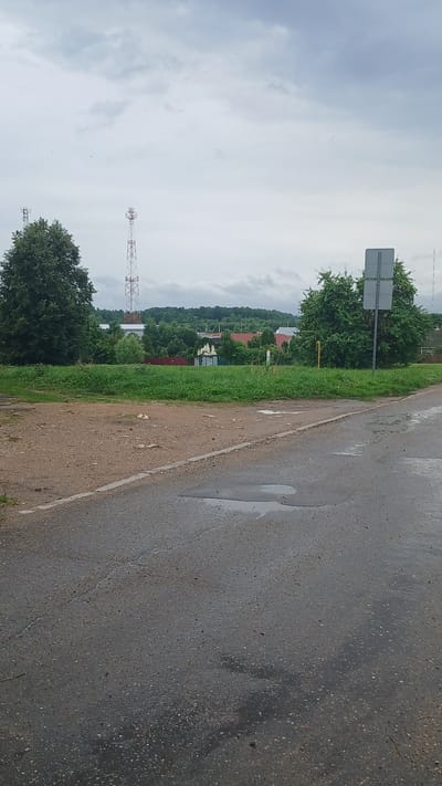 The image depicts a rural or suburban roadside scene under an overcast sky. In the foreground, a wet asphalt road with visible cracks and puddles stretches from the bottom edge towards the right. A dirt or gravel shoulder runs along the left side of the asphalt, separated by a white line. Beyond the shoulder, a grassy embankment leads to a line of trees and buildings in the middle ground. Two tall telecommunication towers, one red and white lattice, and another with antennas, are visible in the distance. A traffic sign with two grey panels stands near a large, leafy green tree on the right side of the middle ground. The background consists of rolling hills covered in lush green trees, with glimpses of red-roofed houses nestled amongst them. The overall atmosphere is calm and depicts a typical day in a non-urban environment, possibly in the vicinity of Ivakino, Russia, given the context. No people or animals are visible in the frame.