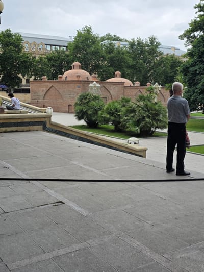 The image depicts a public square in Ganja, Azerbaijan, featuring historical architecture and greenery. In the foreground, a man with short grey hair, wearing a grey patterned t-shirt and black trousers, stands with his back to the camera, holding a bag in his right hand. To the left, on a tiered staircase, a young person with dark hair, wearing a white t-shirt with a number "1" on the back, sits facing away from the camera, looking downwards.

Behind the staircase, a brick structure with several domed roofs is visible, characteristic of traditional bathhouses. This bathhouse is surrounded by lush green trees and palm bushes. In the background, a modern building with a grey roof and windows, displaying signage with the letters "L&GO HOTEL", is partially obscured by trees. The overall environment suggests a warm day with an overcast sky. The ground is paved with large grey stone tiles, and decorative patterned tiles line the staircase.