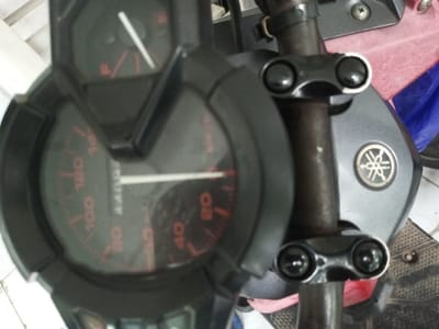 A close-up shot features the speedometer and handlebars of a Yamaha motorcycle. The speedometer, with a black surround and red numbers, displays a speed of approximately 4400 km/h. A fuel gauge is visible above. The rusty handlebars are secured by black clamps with shiny bolts. The Yamaha emblem is present on the motorcycle's body behind the handlebars. The surrounding environment is slightly out of focus, with glimpses of other parts of the vehicle and the floor.