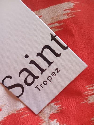A white tag with embossed text is placed on a red and white patterned fabric. The text on the tag reads "Saint Tropez". The tag is angled, with the top left corner towards the viewer. The fabric has a textured, woven appearance. The image does not contain any information about Monsummano Terme, Italy.