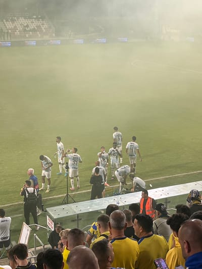 The image depicts a football (soccer) stadium in Nof HaGalil, Israel, heavily obscured by dense, yellowish-white smoke or haze emanating across the green turf. Approximately a dozen football players, primarily in light-colored (white/light blue) jerseys with "Amy Cargo" sponsorship and numbers (e.g., 6, 13, 34), are visible on the field near the sideline. Several players are standing, with at least one (number 34) drinking from a plastic water bottle. Another player (number 21) is bent over, appearing to attend to an object or individual on the ground.

Non-player personnel are also present on the sideline, including an individual in a black shirt standing near a tripod-mounted device, and an older man with white hair near player number 13. LED advertising boards displaying "BLU" are visible along the perimeter, partially obscured by the smoke. Empty stadium seating with red and white seats is discernible in the upper background. A "Winner" sign is partially visible in the far background.

In the immediate foreground, a crowd of spectators is visible from an elevated perspective, primarily showing the backs and tops of their heads. Many wear yellow shirts, indicative of team support. An individual in the foreground wears a head covering featuring a Star of David. A person in an orange safety vest is also visible among the spectators. The scene appears to capture a break in play or pre/post-match activity under conditions of reduced visibility due to pyrotechnic smoke or fog.