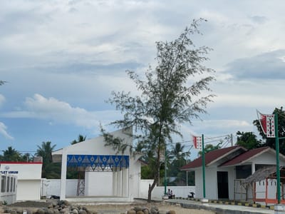 The media file captures an outdoor scene in Lancok, Indonesia, likely during daylight hours with overcast skies and diffused light. The setting appears to be a developing or newly constructed public area, possibly a community center or a rest stop.
Prominently featured is a stark white building with a geometric blue and white decorative frieze. To the left, another section of a white building is visible with the text "'LET UMUM" on its side. This building has a red trim along its roofline. A tall, spindly tree with sparse foliage stands in front of the main white building, its branches reaching towards the cloudy sky.
In the mid-ground, a smaller white building with a red tiled roof is situated to the right. It has a dark entrance and a small, thatched-roof shelter attached to its side. Two flagpoles with distinctive red and white flags that have a stylized bird motif are planted in front of this building. A motorcycle is parked near the buildings, and two people, one appearing to be on a scooter and another person seated, are visible in front of the smaller building.
The ground in the foreground is covered with scattered rocks and gravel, suggesting an unpaved or recently cleared area. Palm trees are visible in the background, indicating a tropical or subtropical environment. The overall impression is one of a simple, functional public space in a coastal or island region. The weather appears calm, with soft light and a muted color palette due to the cloudy conditions.
