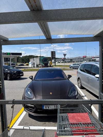 In Berģi, Latvia, a brown Porsche Panamera is parked in a lot under a covered structure, possibly a gas station canopy. The car's license plate reads "LV VV - 19". In front of the Porsche, a metal shopping cart with a red plastic seat is visible, secured by a chain attached to a metal railing. Other vehicles, including a silver minivan, a black SUV, and a green truck, are also present in the parking lot, along with a gas station pump and sign. The sky is blue with scattered clouds, and trees are visible in the background.