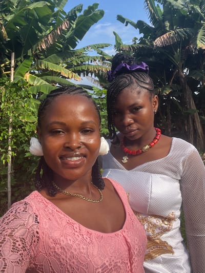 This outdoor portrait features two young Black women posing against a backdrop of lush green foliage and a clear blue sky, likely in Aghalokpe, Nigeria. The scene is brightly lit by natural sunlight, indicating daytime.

In the foreground, a woman with braided hair smiles warmly at the camera. She wears a pink lace top with long sleeves and a prominent gold chain necklace. She has a small stud nose piercing and appears to have light makeup on.

Behind and slightly to her left, the second woman looks directly at the viewer with a serene expression. Her braided hair is pulled back, held by a purple scrunchie. She wears a striking red beaded necklace adorned with a gold cylindrical bead and a small gold charm. Her attire consists of a white garment, possibly a top or dress, featuring a mesh-like texture and elaborate gold embroidery over the chest area.

The background is dominated by large, vibrant green leaves, characteristic of banana plants, with other dense greenery below. The clear blue sky with a few wispy clouds completes the natural setting. The women appear to be enjoying the sunny weather. No visible text is present in the image.