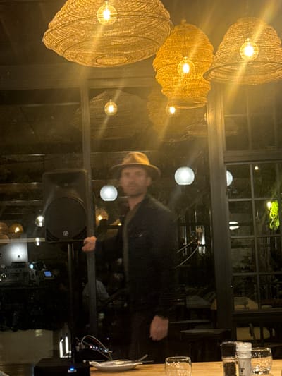 A person with a hat is visible through a glass window or door, with reflections of light fixtures and the surrounding environment visible in the glass. The location appears to be a restaurant or bar, evidenced by the presence of glasses on a counter to the upper right of the image. The presence of what appears to be a PA speaker suggests the possibility of live music or a sound system. There is insufficient information to definitively ascertain the location within Paarl, South Africa.