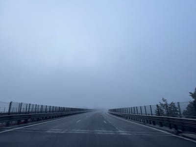 The image shows a foggy highway scene, looking forward along the road. The highway has multiple lanes separated by white lines. On either side of the road are sound barrier fences, some with what appear to be trees or foliage behind them. The sky is a uniform, pale blue-grey, indicating heavy fog or low cloud cover. The road surface appears wet, suggesting recent rain or moisture. There are no people or vehicles visible in the frame. The location is identified as Krushovitsa, Bulgaria.
