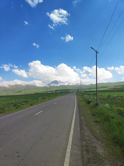 The image shows a rural road in Geghadzor, Armenia, on a clear day with a bright blue sky and white clouds. The road, paved with asphalt, has a white line marking the lanes. To the right of the road, there is a grassy verge with a wooden utility pole supporting electrical wires and a street lamp. The landscape features rolling green hills in the mid-ground, with snow-capped mountains in the distance. A small village with houses is visible nestled in the valley. The foreground is dominated by the road and the grassy area to its side, with some patches of wildflowers. The scene conveys a sense of peaceful, open country.