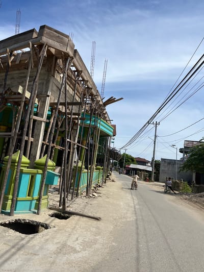 The image shows an outdoor scene in an Indonesian village with a building under construction and a street. The building, painted in teal and lime green, is partially visible, with scaffolding made of wooden poles supporting its structure. The main building material appears to be concrete and wood. The street in front of the building is paved with asphalt and shows signs of wear, with a large hole in the surface near the building.

Two individuals are present on the street. One person is riding a bicycle, and another is walking. The sky is blue with some wispy clouds, suggesting it is daytime. Overhead, power lines stretch across the frame, supported by utility poles. The overall impression is of a developing area, with ongoing construction and daily life unfolding on the street. The time of day appears to be late morning or early afternoon, given the bright sunlight casting shadows. There is no visible text in the image.