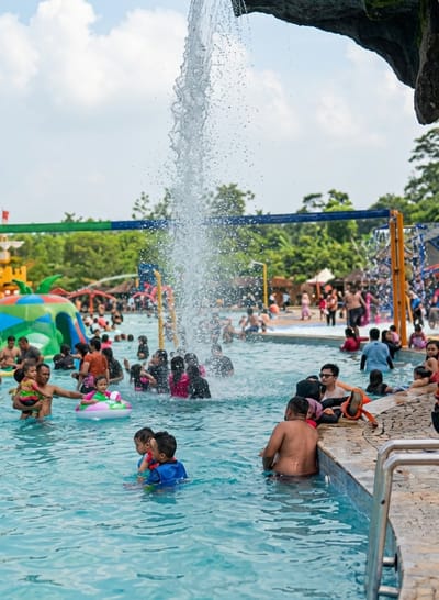 The image shows a crowded outdoor swimming pool at the Pandang Wisata Simbelang waterpark in Durin Simbelang, Indonesia. A man is holding a child near an inflatable donut. Water cascades from a manufactured rock structure above the pool, creating a splash zone amidst the people. In the background, there is green vegetation and various waterslides and playground structures. People of varying ages and ethnicities are scattered throughout the pool, swimming and interacting. Poolside, people are seated or standing near the edge, with towels and other personal items present. A metal handrail is visible in the lower right corner of the frame.