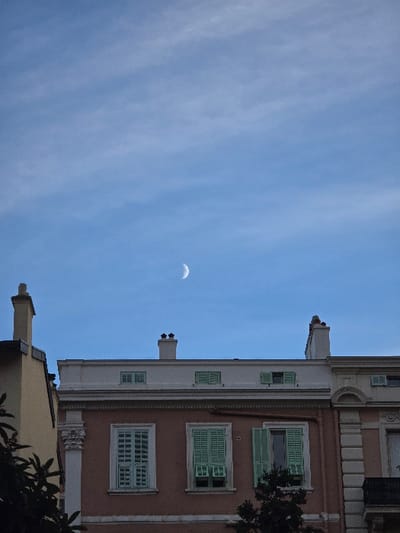 A crescent moon hangs in a pale blue sky. The facade of a building rises on the right side of the frame. Shutters, painted green, adorn the windows set into a reddish-pink wall. A section of a tan-colored building with a chimney is visible at the bottom. Dark foliage peaks in from the right edge of the image. The photo is tilted ninety degrees clockwise.