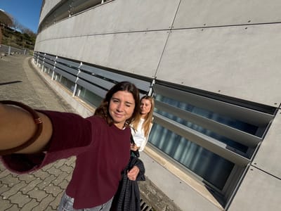 The image depicts two adult females in an outdoor setting in Porto Salvo, Portugal. The subject in the immediate foreground, with brown, wavy hair, wears a maroon long-sleeved top and light-colored trousers. Her left arm is extended towards the camera, suggesting a selfie action, and she exhibits a slight smile. A brown bracelet with copper-colored accents is visible on her left wrist. The second subject, positioned behind and to the right, has long, blonde hair and wears a white long-sleeved top. She carries a dark, patterned jacket over her arm and faces slightly to her right, with her mouth partially open.

Both individuals stand on a paver block surface composed of grey-brown interlocking concrete blocks. A metal drainage grate is visible on the pavement near the building's base. The prominent background feature is a modern, curved building with a light grey paneled facade, marked by visible horizontal and vertical seams and rivets. Along the lower section of the building, a horizontal strip of windows is shielded by angled grey external sun louvers. In the far left background, a vegetated hillside with bare trees is visible. The scene is brightly illuminated by direct sunlight under a clear blue sky, indicating daytime conditions.