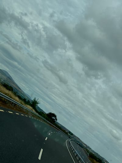 This is a tilted, eye-level, medium shot of a highway in the New Ross Municipal District, Ireland. The sky is overcast with thick, grey clouds that dominate the upper portion of the frame. A mountain range is visible in the distance on the left side of the image, partially obscured by the clouds. The highway is paved with asphalt and marked with white lines, including a dashed white line to the left of the lane and a solid white line to the right. The road curves to the right, with a guardrail separating it from a grassy embankment. Some dry grass and a few trees are visible on the embankment. The scene is likely captured from a moving vehicle, given the perspective and the slight blur in some areas. There are no people or animals visible in the image.