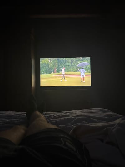 A person rests in bed, a blanket beside them. A television screen displays a golf scene, showing a golfer swinging their club on a green, with a caddy holding an umbrella nearby. The room is dimly lit, with the screen providing the main source of light.