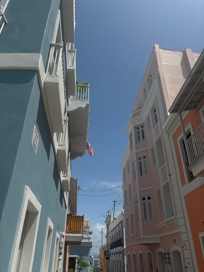 The image depicts a narrow street in Old San Juan, Puerto Rico, USA, with colorful colonial-era buildings on either side. The building on the left is painted a vibrant teal and features white trim and balconies. A street sign on this building reads "CALLE DE LA TANCA". On the right, buildings are painted in shades of pink and orange, also adorned with white trim and balconies. A Puerto Rican flag is visible hanging from a balcony. The sky is a clear, bright blue with a few wispy clouds. Electrical wires are strung across the sky, and a distant view of other buildings and greenery can be seen further down the street. The architecture suggests a historic urban environment, typical of Old San Juan.