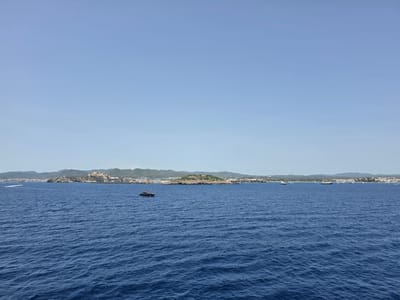The image captures a wide view of the sea off the coast of Santa Eulària des Riu, Spain, under a clear, light blue sky. The foreground is dominated by the deep blue, rippling water. In the mid-ground, several boats are visible. A prominent dark-colored motor yacht with a red accent is positioned to the left of center. Further to the right, a line of white sailboats and yachts are anchored or moving slowly along the coastline. The background reveals the city of Santa Eulària des Riu, characterized by white buildings and structures nestled against rolling green hills. A notable feature on a prominent outcrop is a fortified building, likely a castle or a historical structure, overlooking the sea. The overall scene is serene and depicts a coastal Mediterranean landscape.