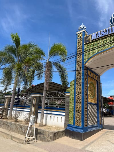 The image depicts the entrance archway to a mosque in the city of Dalam, Indonesia. The archway is adorned with intricate gold and green geometric patterns against a blue background. The word "MASJID" is prominently displayed in blue lettering above the arch. Flanking the arch are palm trees and a white wall with blue and gold accents. In the foreground, a stone wall borders a pathway. The sky above is clear and blue, with a few wispy clouds. The scene appears to be a sunny day in a tropical environment.