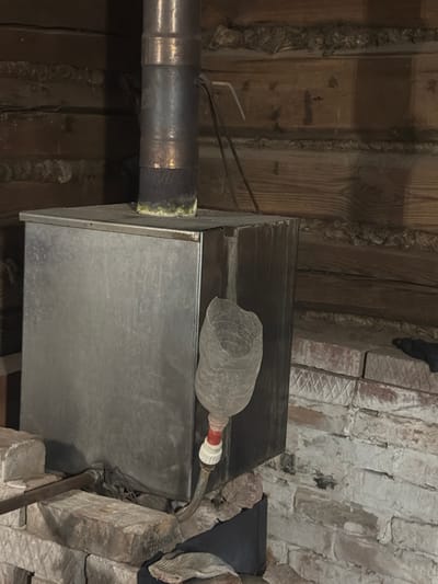 The image shows a close-up, low-angle view of exposed HVAC ductwork and surrounding infrastructure, likely in a basement or crawl space. The primary objects are a large rectangular metal duct, a smaller circular metal pipe running horizontally to the left, and what appears to be a plastic bottle attached to the circular pipe with tape and a hose clamp. The bottle is filled with a gray, lumpy material and has a red and white connector.
The setting is characterized by rough, unfinished materials. The ceiling consists of rough-hewn wooden beams and planks, some of which appear weathered or aged. To the right, a brick wall with a rough, uneven surface is visible, with some decorative or structural elements suggesting an older construction style. There are also some metal conduits and wires visible, indicating the presence of electrical or plumbing systems. The overall impression is one of utilitarian, possibly older, construction, consistent with a service area or utility space.
No people or specific activities are depicted. The lighting is dim, suggesting an indoor environment, possibly without active artificial light sources, or the image was taken with a flash. There is no discernible text in the image. The scene is devoid of any strong emotional cues, presenting a purely observational documentation of the infrastructure. The location context "Аргамак, Russia" provides a geographical reference but no visual cues within the image directly confirm this location.