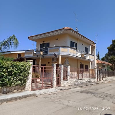 The image shows a two-story house with a light yellow facade, red tile roof, and a balcony on the second floor. The house is surrounded by a stone and metal fence. A lush green hedge is visible along the fence on the left side of the image. A palm tree is also present. The house has several windows with dark shutters. An air conditioning unit and a solar panel are mounted on the exterior wall. The scene appears to be a residential street in Grumo Appula, Italy, with a clear blue sky. The timestamp "16.07.2026 14:27" is overlaid on the bottom right of the image. There are no people visible in the image.