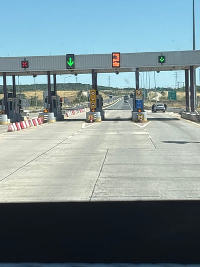 The image depicts a toll plaza on a sunny day, likely on a highway leading to or from the city of Ardani, Greece, given the context of the provided crops. The perspective is from a vehicle approaching the plaza. Several toll booths are visible, with overhead signs indicating lane status. A red 'X' is displayed above one lane, signifying closure. Another lane shows a green downward arrow, indicating it is open. A third lane displays an orange sign with what appears to be a number '2' and an arrow.
Various traffic control elements are present, including red and white striped barriers delineating lanes and traffic cones. Signal lights are visible on some of the toll booth structures, with at least one amber light illuminated. Several cars are in the process of navigating the toll plaza, with one white car stopped at a booth and a person standing next to its open driver-side door. In the background, power lines and vegetation are visible under a clear blue sky. The road surface is asphalt with white lane markings.