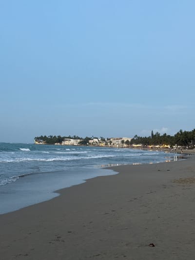 The image shows a beach scene in Cabarete, Dominican Republic. The sky is blue with a few light clouds. The beach is composed of light tan sand. The ocean is visible with waves gently rolling onto the shore. Along the coastline, there are buildings and palm trees, likely hotels or resorts. The overall scene gives a peaceful and serene impression of a tropical beach.