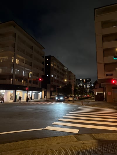 The image depicts a nighttime street scene in Chiba, Japan, featuring an intersection with a pedestrian crossing. To the left, a multi-story building with balconies and illuminated windows dominates the frame. Across the intersection, another building complex stretches into the distance, also showing lit windows. A traffic light, displaying a red signal, is visible on a pole near the right side of the image. Two individuals are walking on the sidewalk in the middle ground. Several vehicles are present, including cars on the road and one parked on the left side of the street. The sky is dark and overcast. The foreground shows a portion of the road and sidewalk with tactile paving.