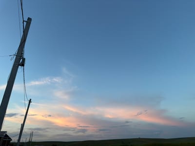 The image depicts a rural landscape under a twilight sky. A utility pole, tilted and with wires attached, stands prominently on the left side of the frame. In the distance, a line of similar poles stretches across a rolling, green terrain. The sky is a gradient of soft blue, transitioning into hues of pink and orange where clouds are illuminated by the setting sun. The horizon line is defined by the dark silhouette of hills and fields, with a small, dark structure visible on the far left at the base of the main pole. The overall scene suggests a peaceful, early evening in a countryside setting, possibly in the vicinity of Нижний Суык-Су, Russia.