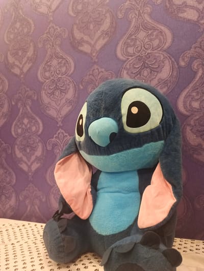 A plush toy resembling the character Stitch from Disney's Lilo & Stitch franchise is seated on a white patterned surface, possibly a bed. The toy is predominantly dark blue with lighter blue accents on its chest and nose. It has large, light blue eyes with black pupils and white highlights, and long, pink-lined ears. The background is a purple wallpaper with a damask-like pattern. The image is cropped to focus on the toy and its immediate surroundings. The scene does not provide any information to determine its location in Putilkovo, Russia.