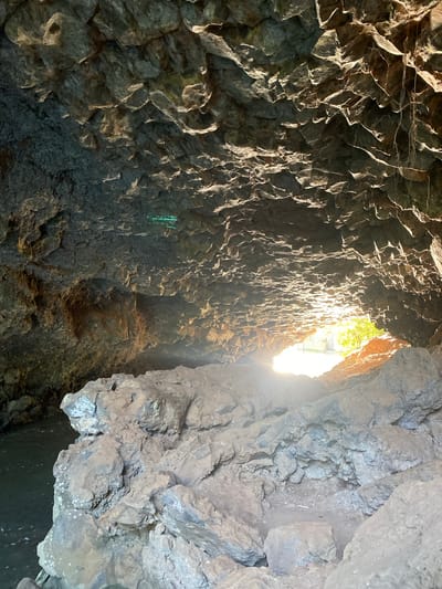The image shows the interior of a cave in Mururi, Kenya. The walls are made of dark, jagged rock. Light pours in from the cave's opening, illuminating the interior. There are no people visible in the image. The environment is rocky and dimly lit.
