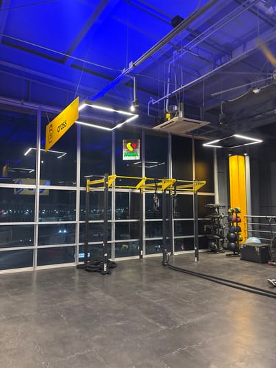 A fitness center with a cross-training area. Yellow and black gym equipment is arranged near a large window. A CrossFit banner hangs near a blue-lit ceiling. Wall-mounted medicine balls and battle ropes are also present. The floor is gray with an interlocking tile pattern. Modern, square LED lighting is visible in the ceiling design. A sign with a green and red logo is placed on the wall.