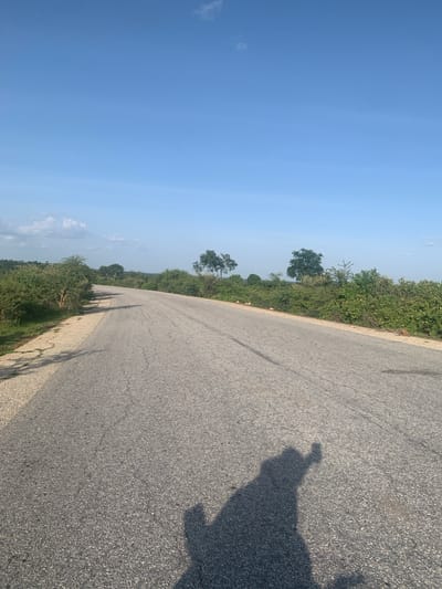 The image shows an asphalt road curving to the right, bordered by lush green vegetation on the left. The sky is a clear, bright blue with a few scattered white clouds. A shadow of a person holding a camera is visible on the road in the foreground, suggesting the photo was taken by someone walking or standing on the road. The lighting indicates it is daytime, likely late morning or early afternoon. The scene appears to be in a rural or semi-rural area, possibly in Nigeria, given the location context. The vegetation is dense and appears to be native to a warm climate. There are no visible signs of text in the image. The overall mood is peaceful and serene, capturing a moment of travel or exploration in a natural landscape.