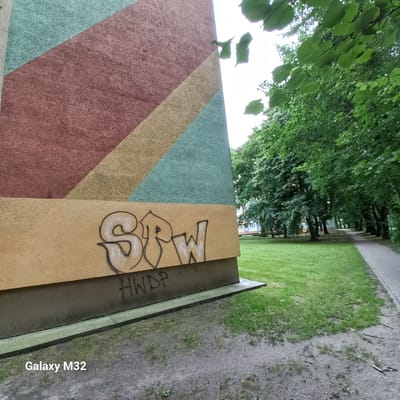 The image shows a section of a building with abstract geometric patterns in shades of pink, yellow, and teal. On the lower, tan-colored section of the building, there is graffiti. The graffiti on the top part of the tan section appears to be in white with black outlines and reads "SPRW". Below this, there is more graffiti in black, reading "HWDP". The building is situated next to a grassy area with trees and a paved walkway. In the background, other buildings can be seen, along with more trees. The scene is outdoors, likely in a residential area of Olsztyn, Poland, given the metadata. The lighting suggests it is daytime, possibly overcast, with visible green foliage from deciduous trees.