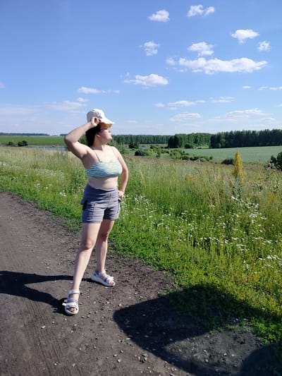 A woman wearing a white bucket hat, glasses, a green and white striped bikini top, and gray shorts stands on a dirt path. She is looking up and to her right, shielding her eyes with her left hand. Her right hand is on her hip. She is wearing white sandals.
The scene is a rural landscape under a bright blue sky with scattered white clouds. To her right is a field of tall green grass and wildflowers, with a treeline in the distance. Behind the treeline, a large green field is visible, possibly a crop field, and a body of water can be seen to the left. The sun is shining brightly, casting a shadow of the woman on the dirt path. The time of day appears to be midday or early afternoon. The overall mood is relaxed and summery. The location context is Блохино, Russia. There is no visible text in the image.