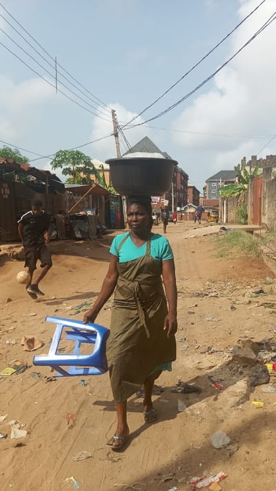 The image depicts a daytime street scene in Awada, Nigeria, under a partially cloudy, light blue sky. The ground is an unpaved, dusty reddish-brown dirt path, littered with various debris including plastic bags, wrappers, and fragments. Shadows indicate a bright sun.

In the immediate foreground, an adult female with dark skin walks forward, slightly right of the center, facing partially left. She wears a light blue short-sleeved top, a dark olive-green apron or wrap skirt, and light-colored flip-flops. On her head, she balances a large, black, wide-rimmed basin containing a white, granular substance. In her right hand, she carries a bright blue plastic stool.

To her left and slightly behind her, a male individual with dark skin, wearing a black long-sleeved athletic shirt with white shoulder accents and black shorts, is actively engaged in kicking a white and black soccer ball. He is mid-stride, moving left to right across the frame.

The midground features a mix of informal structures and more permanent buildings. On the left, behind the soccer player, are several rudimentary stalls or structures constructed from wood, corrugated metal, and other materials. Green foliage is visible behind these structures. On the right, a concrete block wall with a jagged top border runs along the path, partially obscured by large green leaves.

In the background, multiple-story buildings with brick-colored and grey facades and dark roofs line both sides of the street. Distant figures of people are visible further down the path, along with a yellow tricycle. Overhead, multiple black electrical wires crisscross the sky, supported by wooden utility poles.