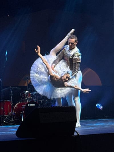 Two ballet dancers in costume perform. A man in a brown and white matador-style outfit supports a woman in a white tutu. The female dancer extends her arm and leg elegantly, while the man balances her. A Yamaha drum set and speaker are visible in the background. The lighting is theatrical, with a spotlight illuminating the dancers and a blue light casting shadows.